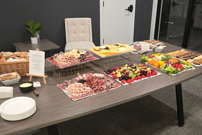 A catering table set up with assorted food platters, cheeses, and drinks in a modern office space. The wall behind features the logo for Advantage Property Management.
