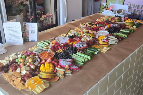 A long countertop covered with assorted packaged snacks, fruit, and small food items arranged in clear plastic bags, photographed in a real indoor setting with a tiled counter and display case visible.